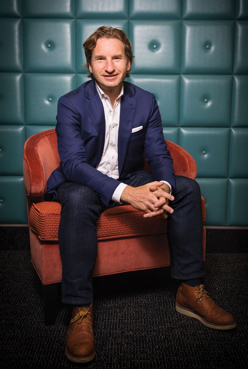 A portrait of Dean Phillips in a suit sitting on a red chair.
