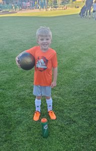 Blonde boy wearing orange shirt and soccer cleats smiling and holding a soccer ball